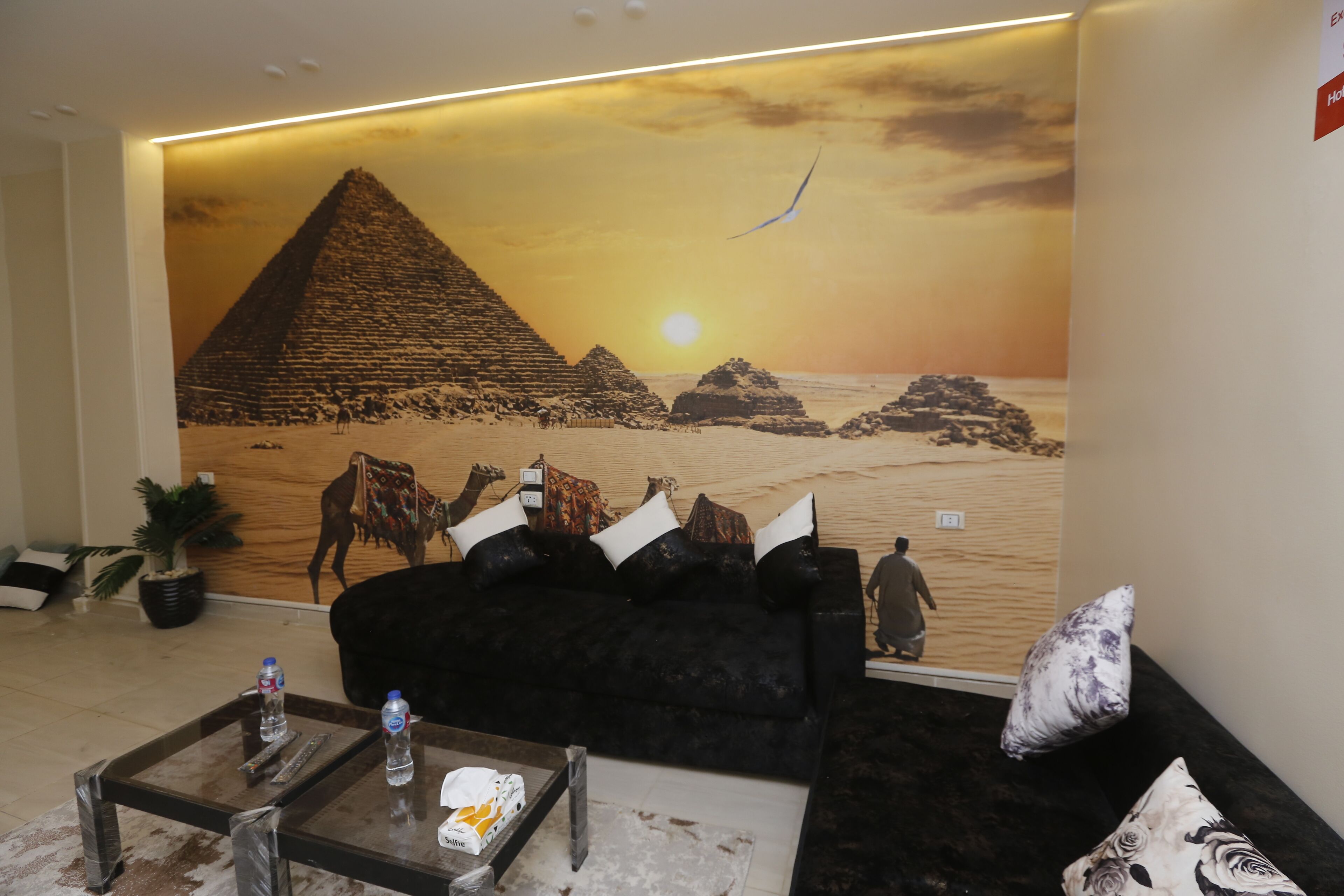 A lobby with a large wall mural of pyramids, a black sofa, a glass coffee table with water bottles, and a potted plant.