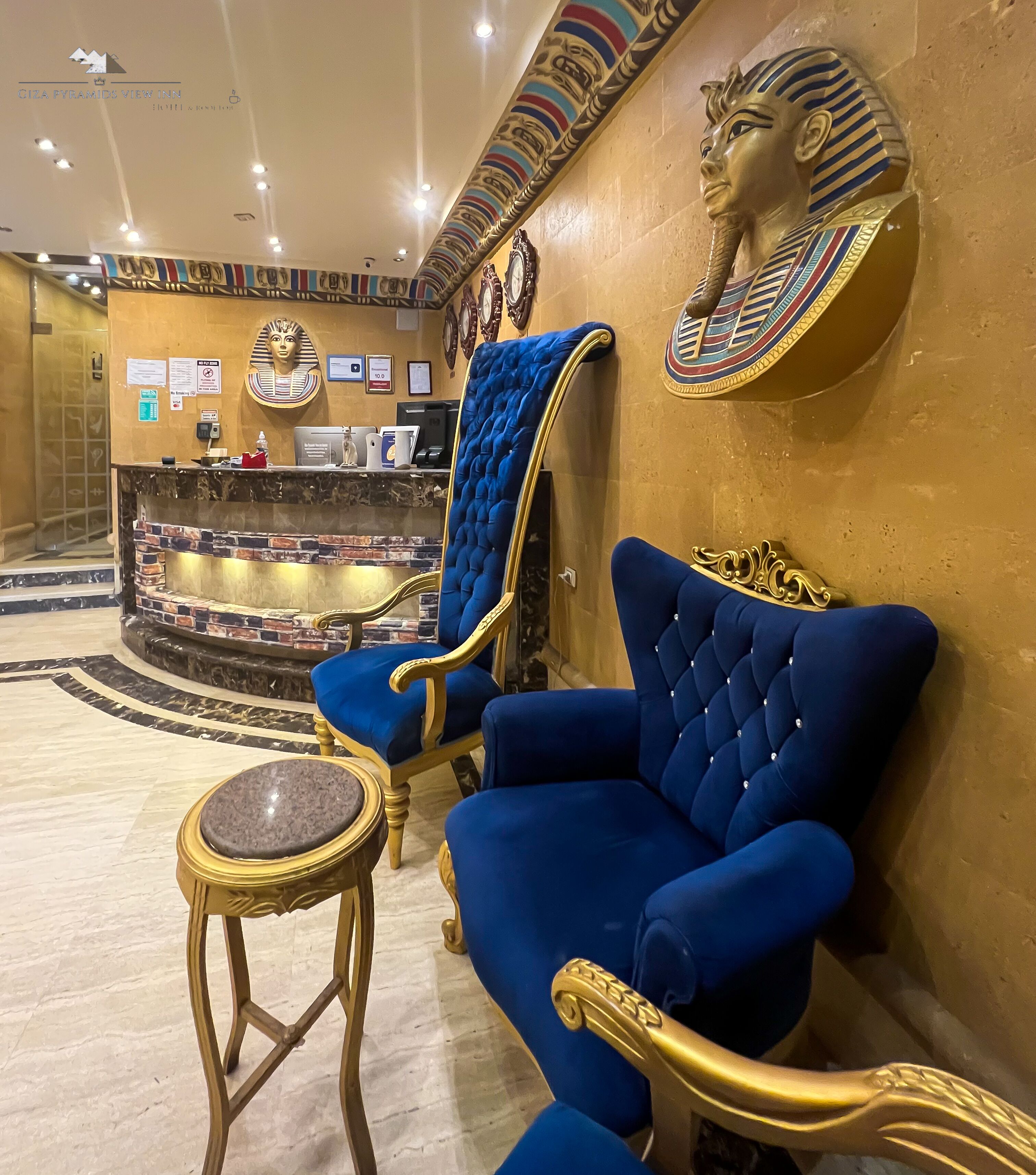 A luxurious lobby with a reception desk, a blue armchair, a gold-framed chair, and an Egyptian-themed wall decoration.