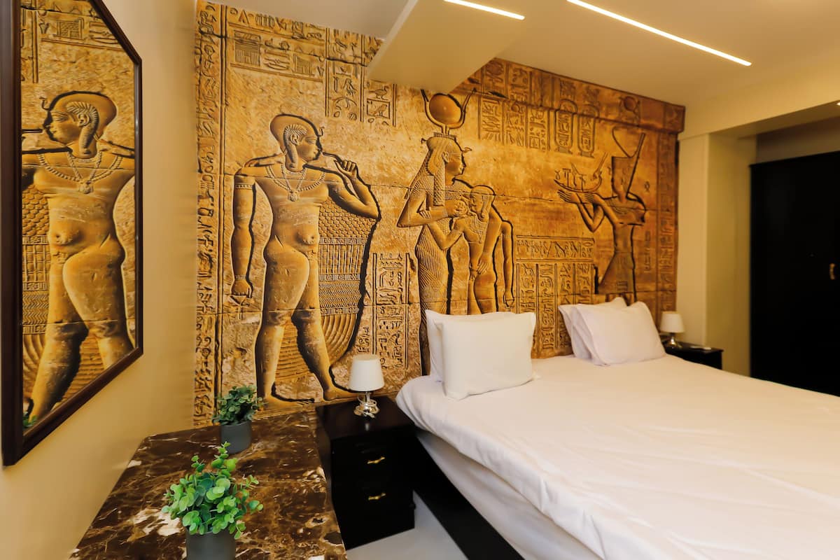 Et hotelværelse med en seng, to natborde, et spejl og en væg med egyptiske vægmalerier.