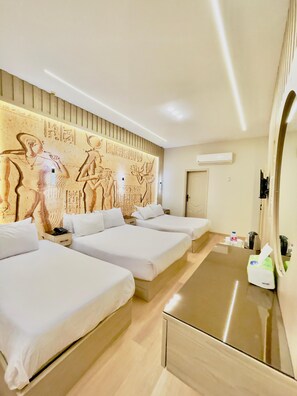 A hotel room with three beds, a wooden headboard with hieroglyphs, a flat-screen TV, and a minibar.