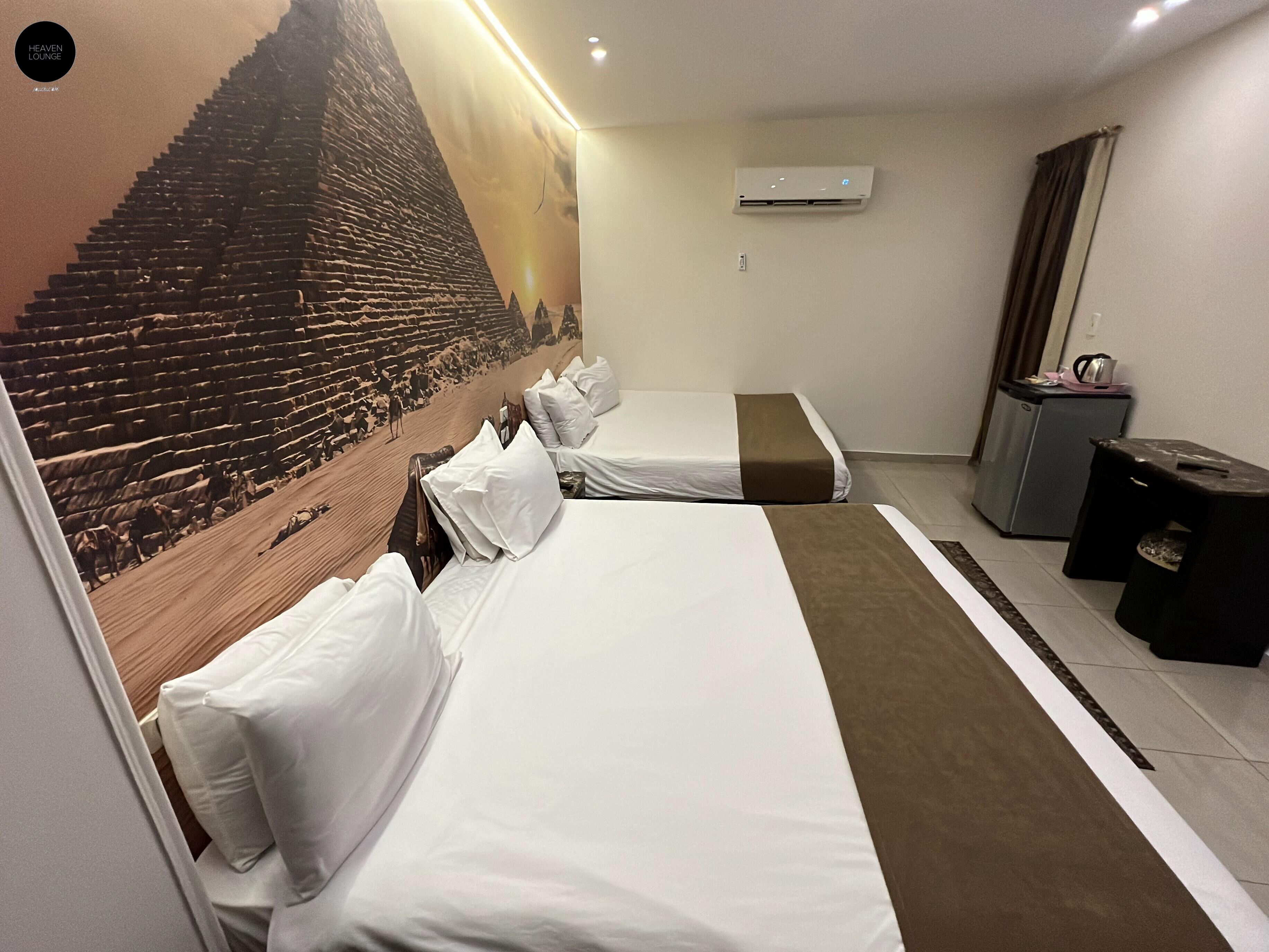 A hotel room with two beds, a wall mural, an air conditioning unit, a small desk, and a trash bin.