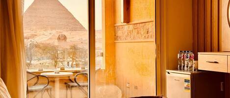A hotel room with a view of the Great Pyramid, a minibar, and a dining area with chairs.