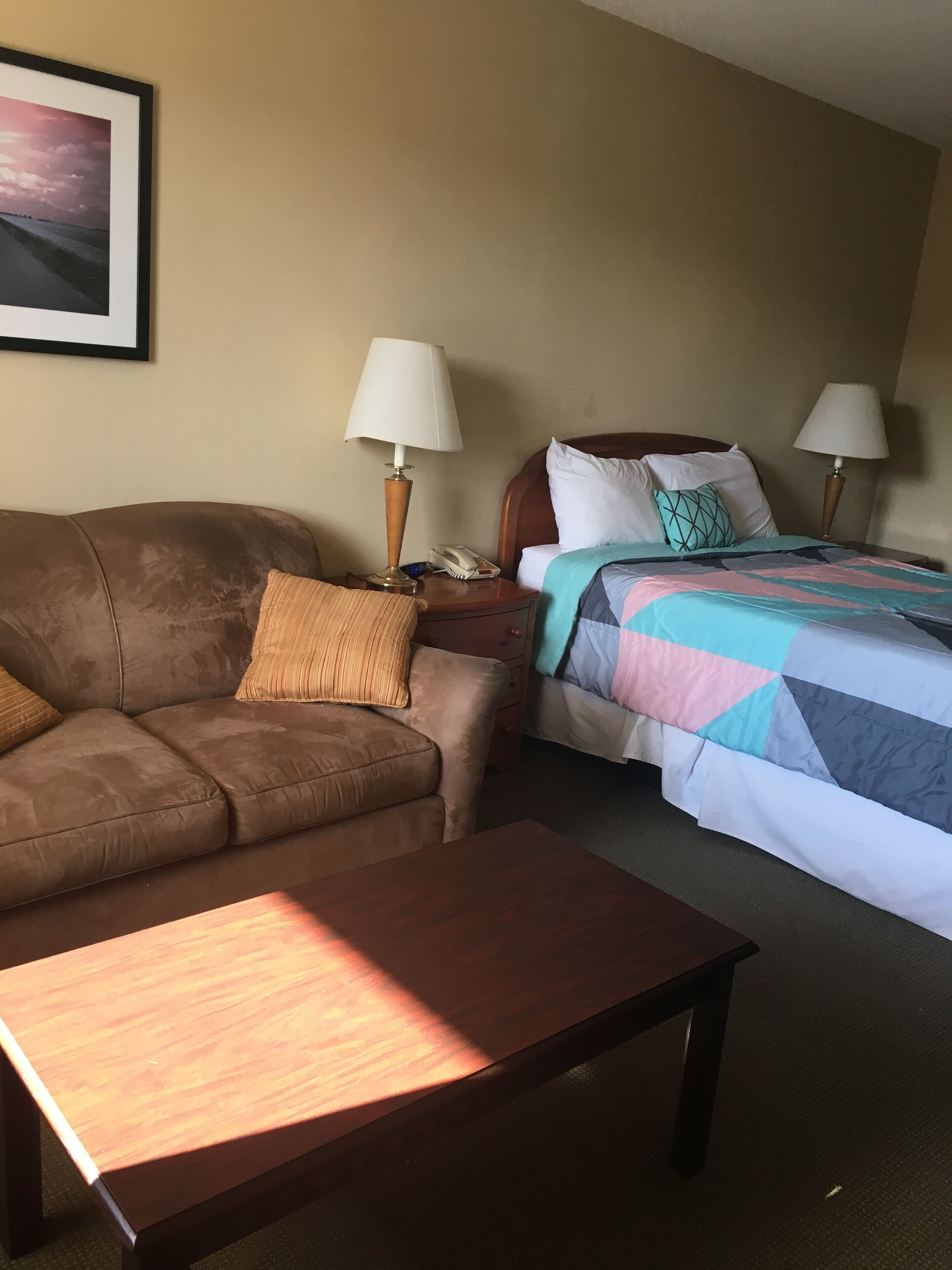 A hotel room with a brown sofa, a wooden coffee table, a bed with a colorful quilt, two bedside lamps, and a framed picture on the wall.