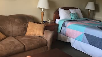 A hotel room with a brown sofa, a wooden coffee table, a bed with a colorful quilt, two bedside lamps, and a framed picture on the wall.