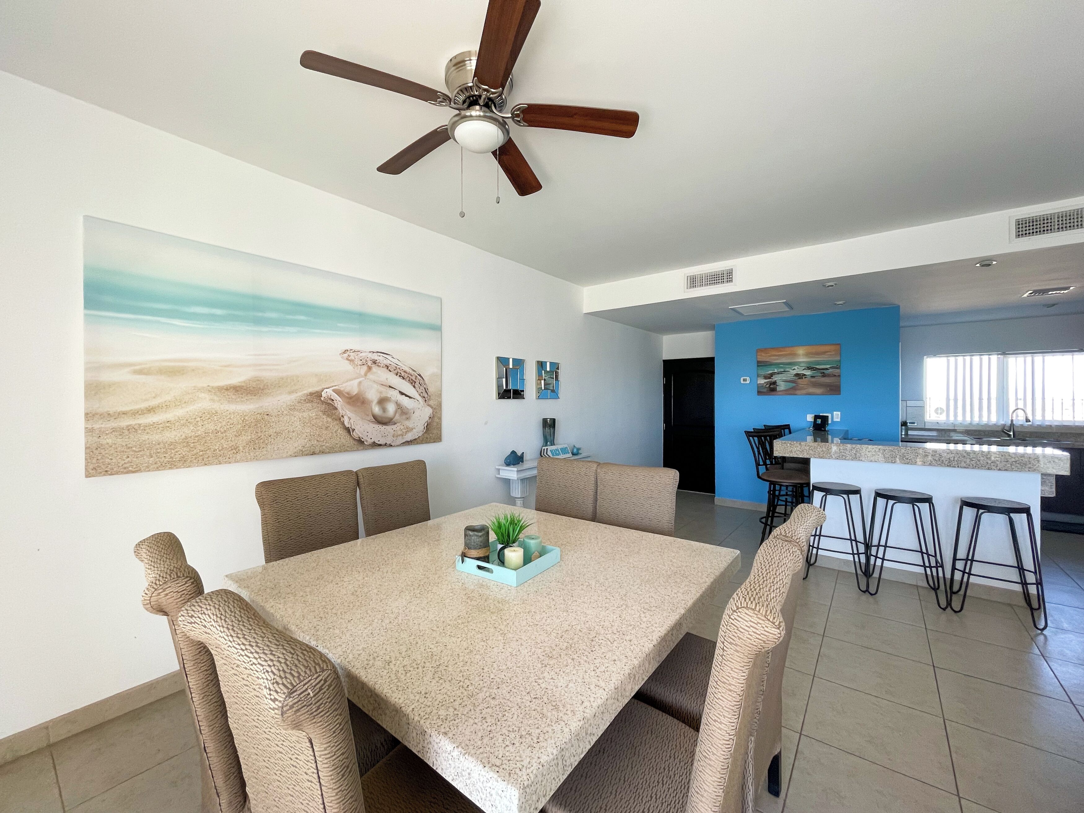 Un comedor con ventilador de techo, un cuadro grande con motivos playeros y una cocina al fondo.