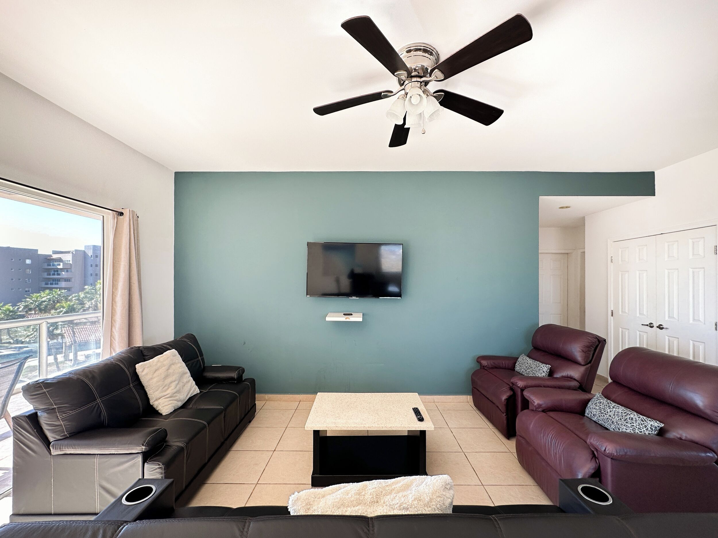 Un salon doté d’un ventilateur de plafond, d’un téléviseur à écran plat, de deux canapés en cuir, d’une table basse et offrant une vue sur les immeubles par une fenêtre.