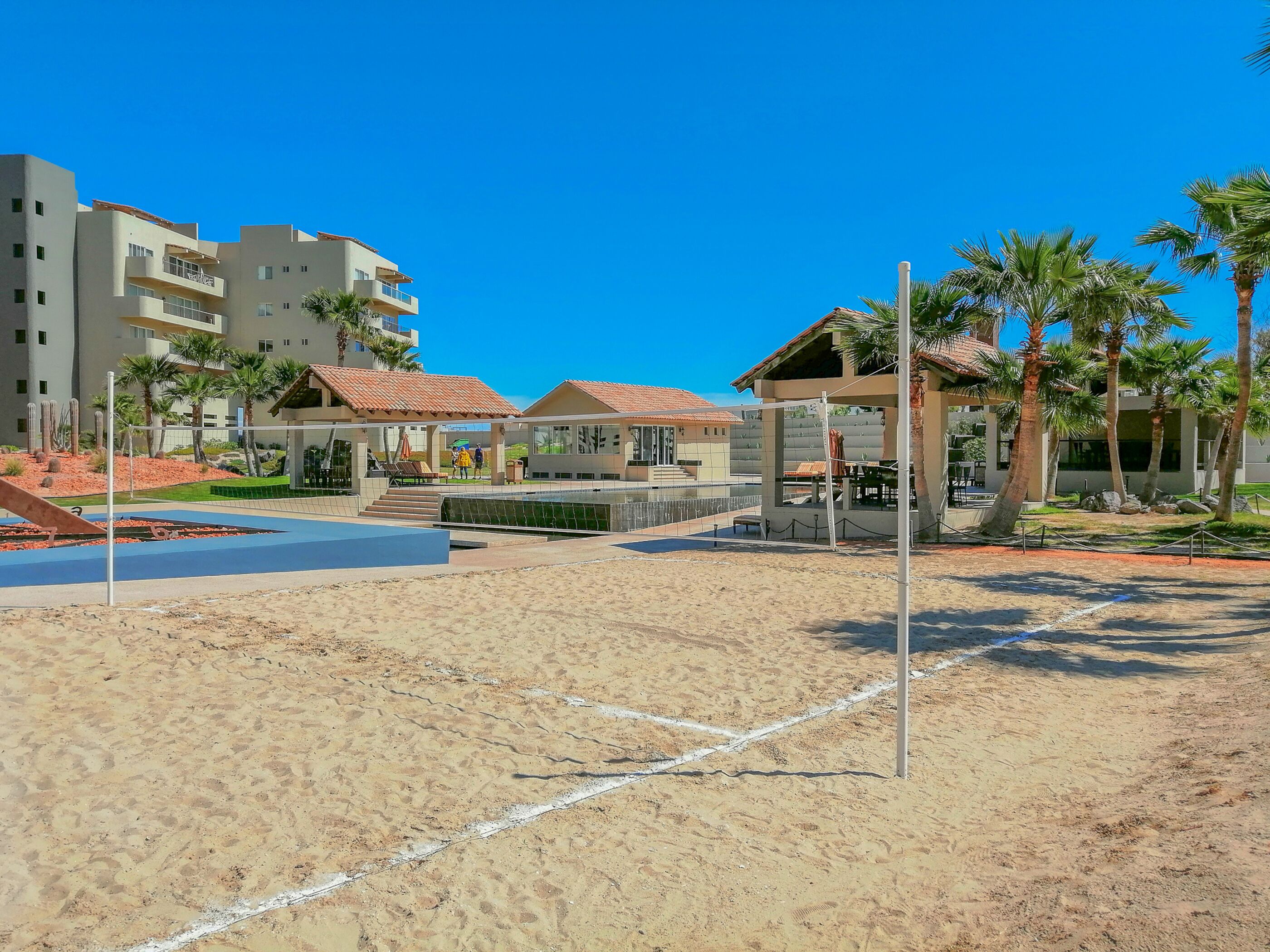 A sandy beach area with a volleyball net, palm trees, and a building with a red-tiled roof.