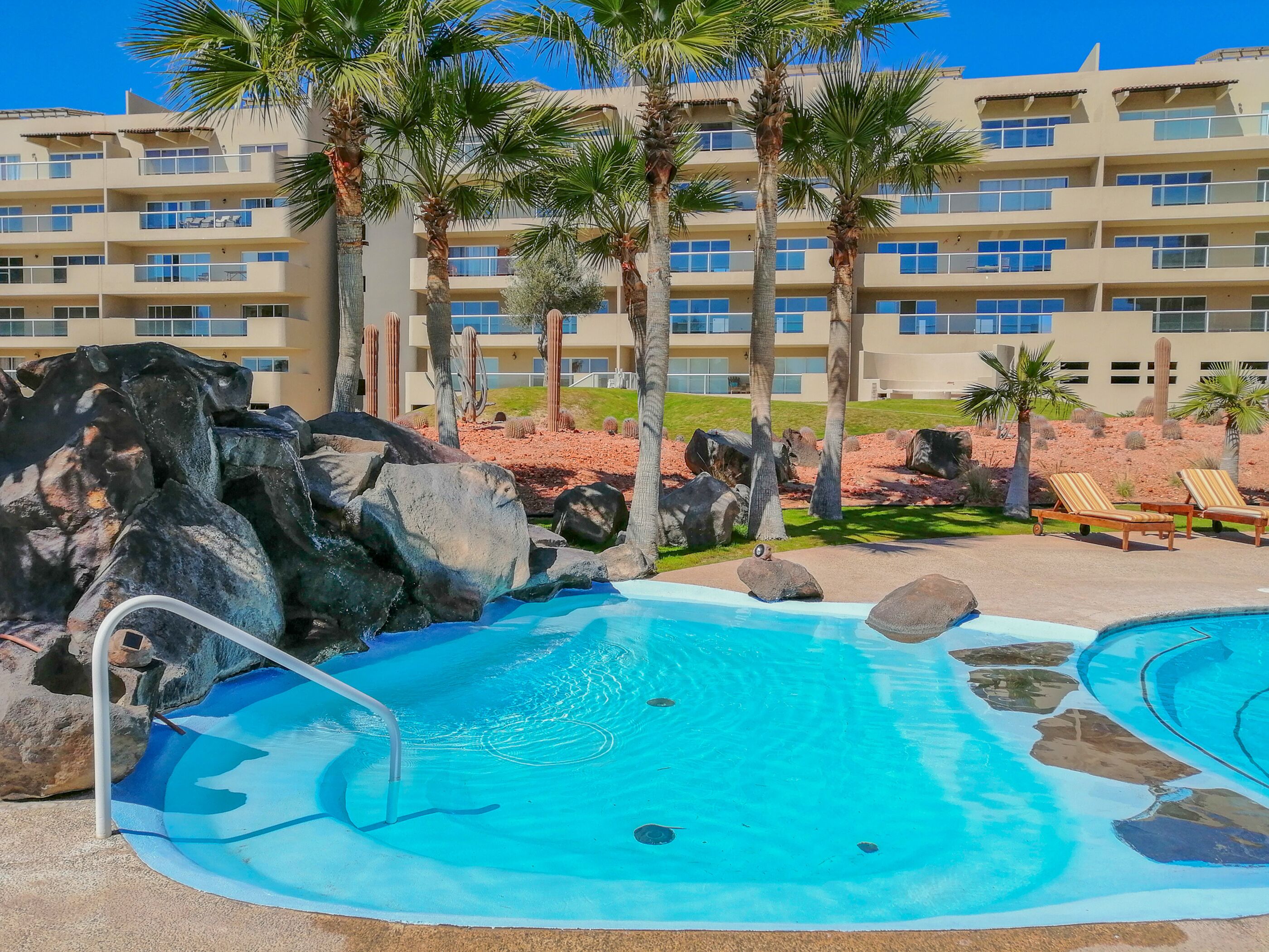 A pool area with a rock feature, palm trees, and a hotel building in the background.