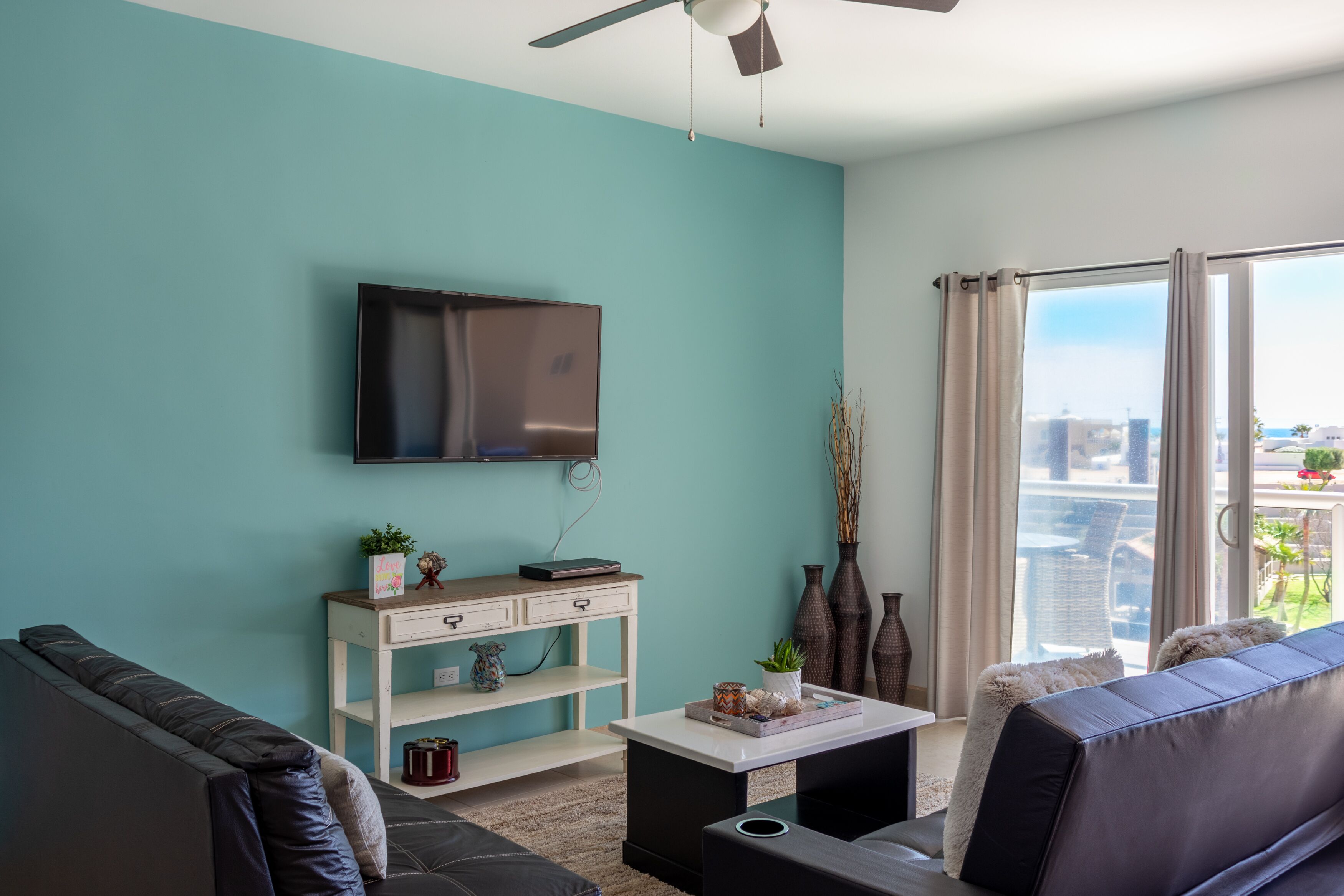 A modern living room with teal walls, a flat-screen TV, a sofa, a coffee table, and a view of the city through large windows.
