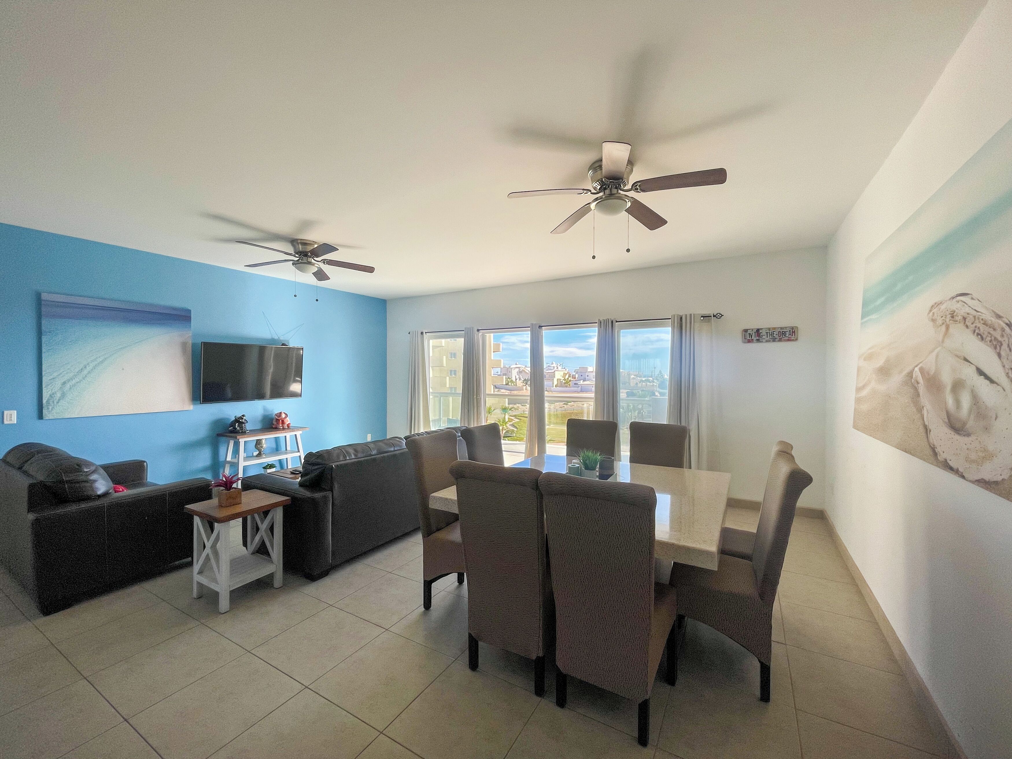 A modern living room with a blue accent wall, a flat-screen TV, a ceiling fan, a dining area with a table and chairs, and large windows offering a view of the city.