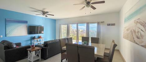 A modern living room with a blue accent wall, a flat-screen TV, a ceiling fan, a dining area with a table and chairs, and large windows offering a view of the city.
