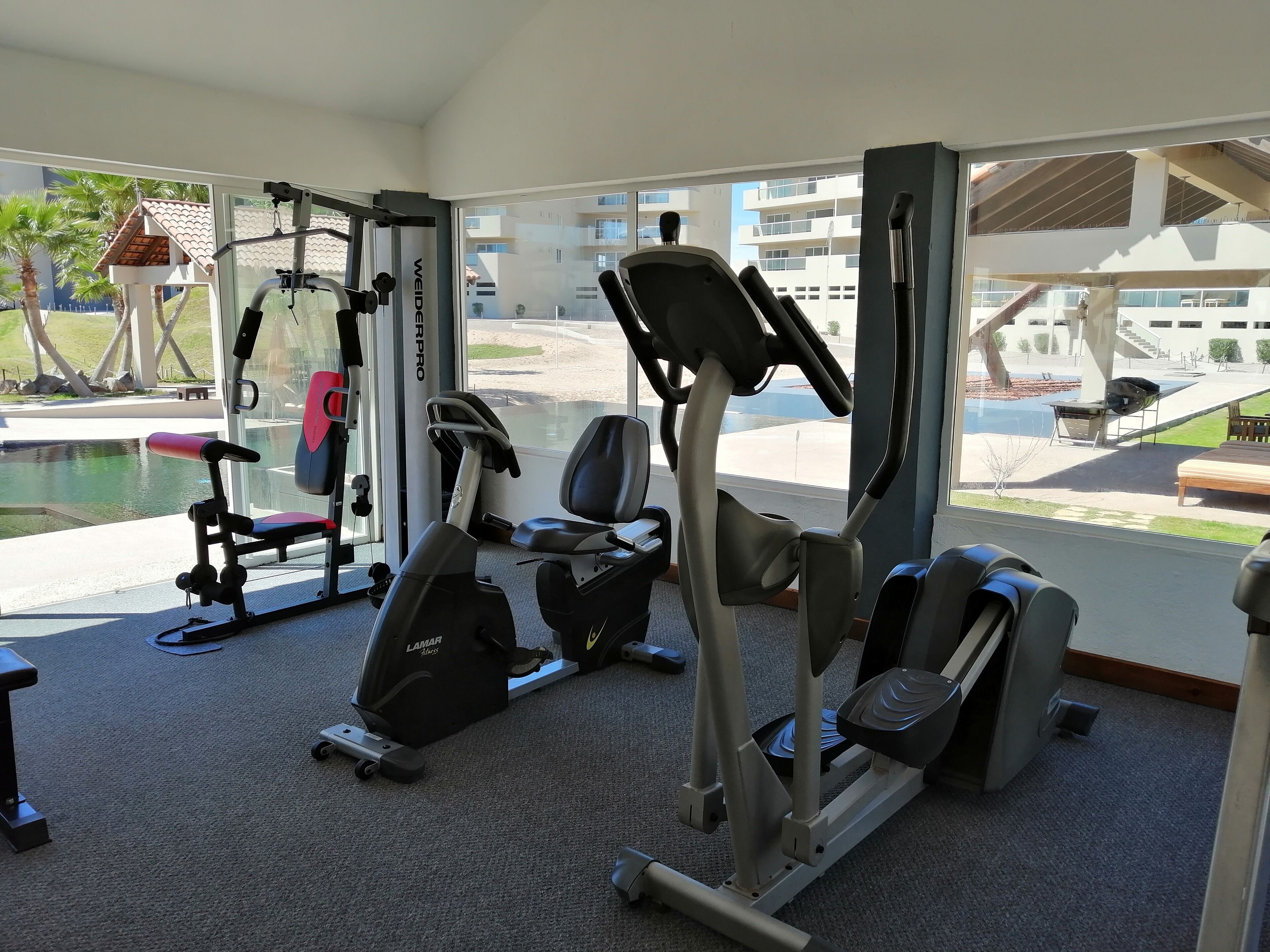 A gym with various exercise equipment, large windows, and a view of an outdoor swimming pool and buildings.