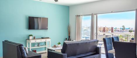 Un saló amb una paret color turquesa, un ventilador de sostre, una televisió de pantalla plana, un sofà, una taula de centre i vista a la ciutat a través de grans finestres.