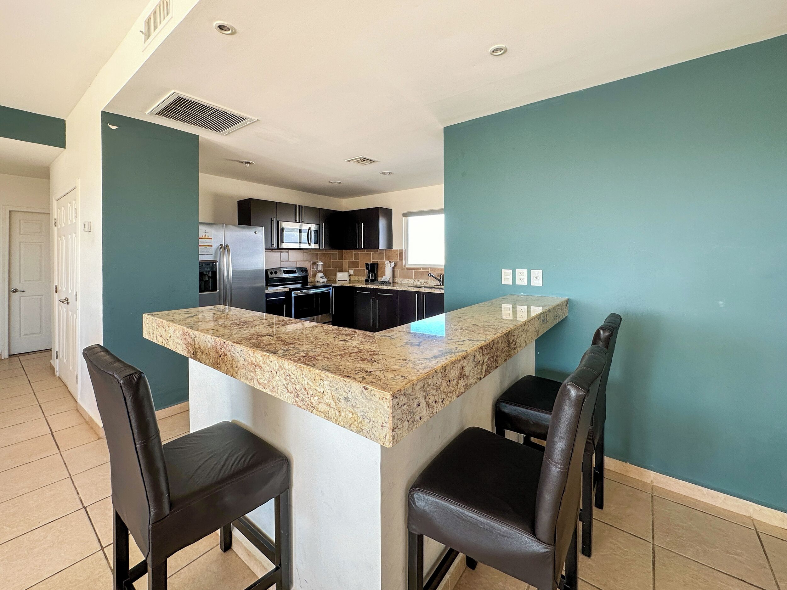 A modern kitchen with a granite countertop, black cabinets, and stainless steel appliances. There are two black leather bar stools and a white door in the background.