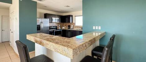 A modern kitchen with a granite countertop, black cabinets, and stainless steel appliances. There are two black leather bar stools and a white door in the background.