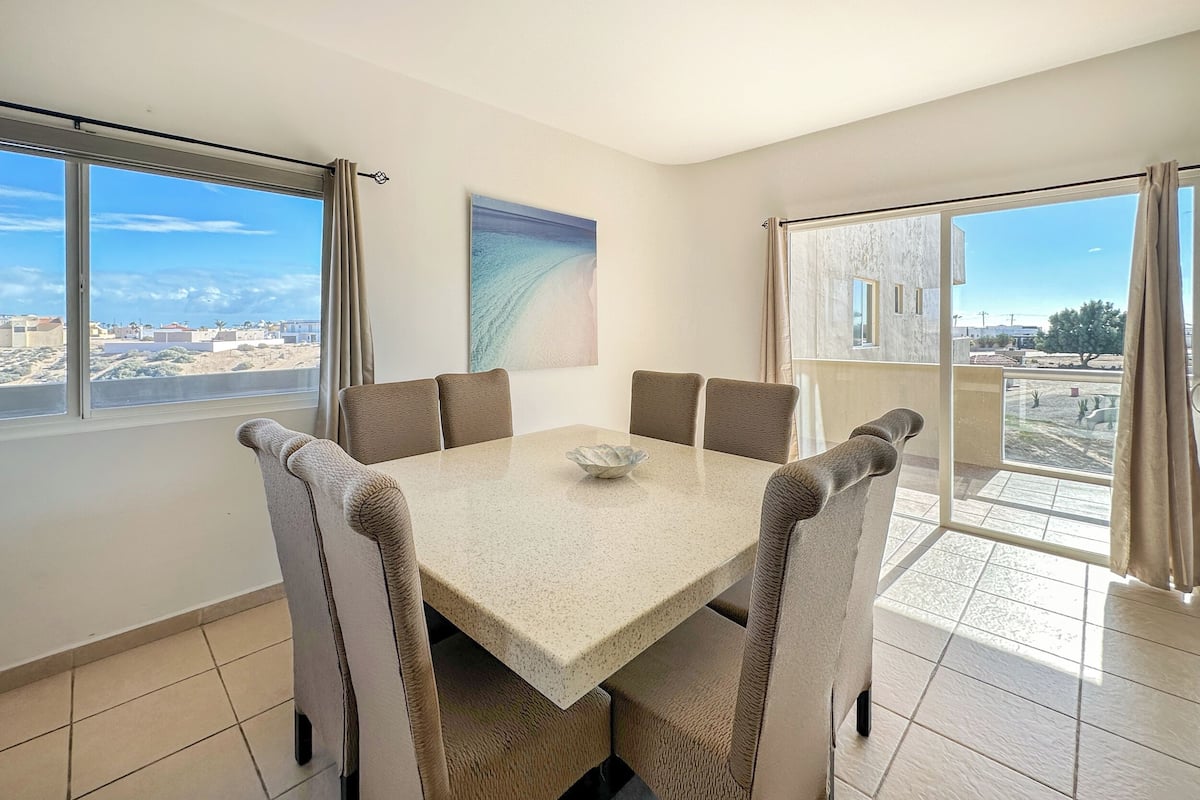 Un comedor con una mesa y sillas, ventanas amplias y un cuadro en la pared.