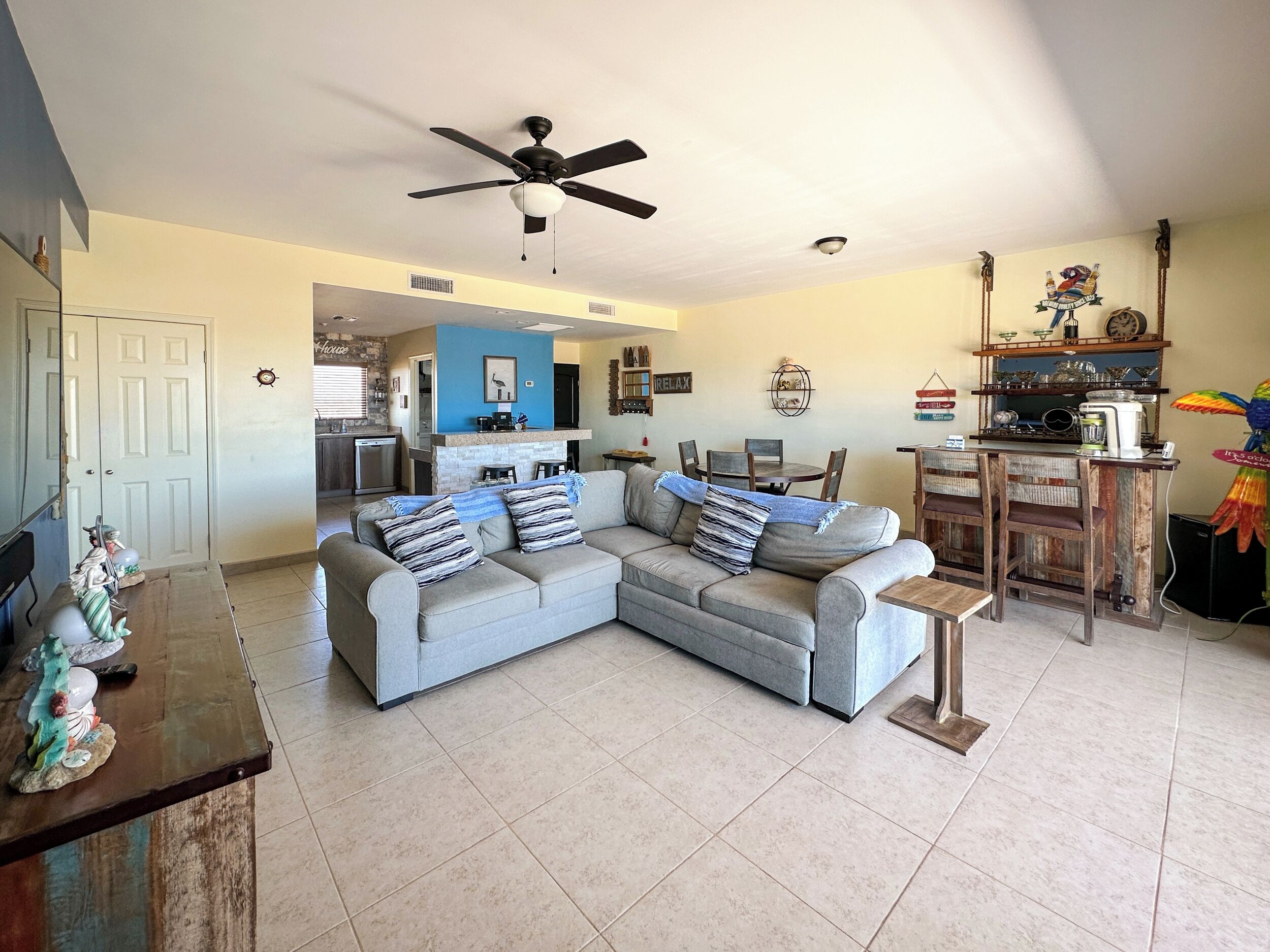 A spacious living room with a grey sectional sofa, a ceiling fan, and a kitchen area in the background.