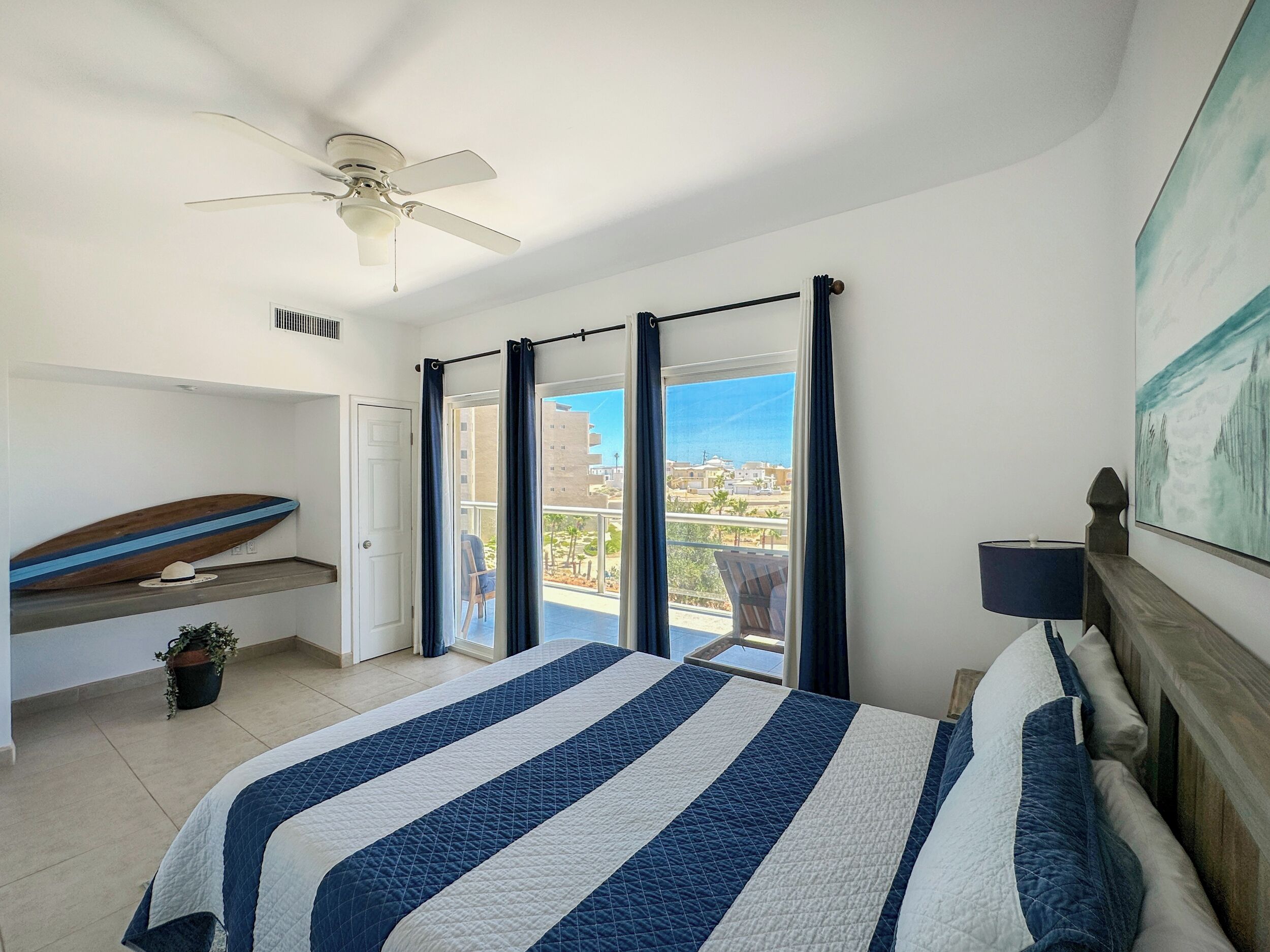 Une chambre avec un lit, un ventilateur de plafond, une vue sur l’extérieur et une planche de surf posée sur une étagère.