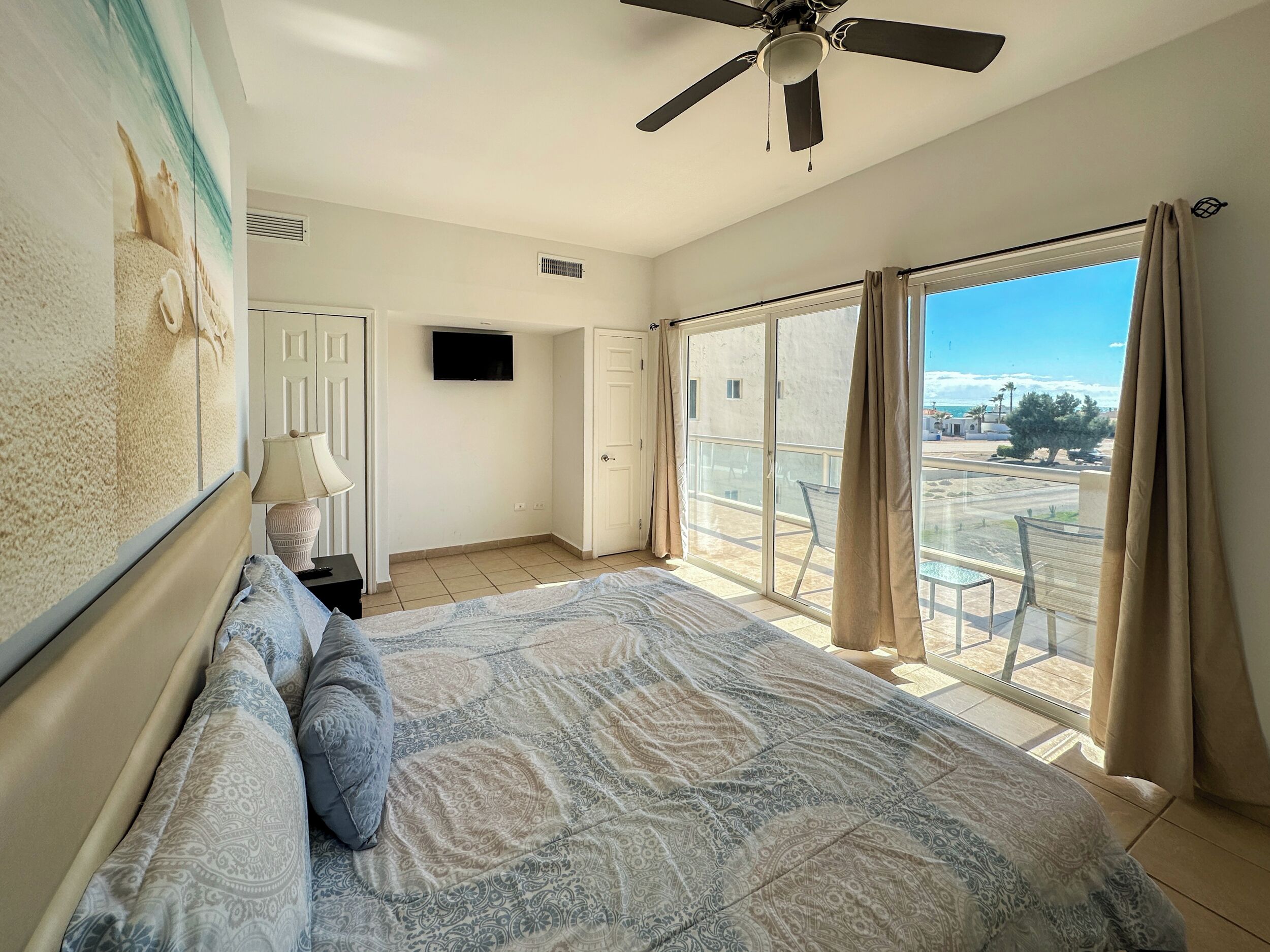 A bedroom with a bed, a ceiling fan, a TV, and a balcony with a view of the beach.