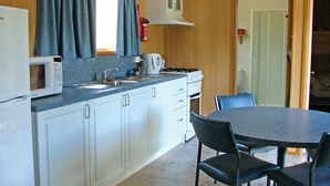 A compact kitchen with a microwave, stove, sink, and cabinets. There is a dining table with chairs and a window with curtains. - Hobart Bush Cabins (Kingston)