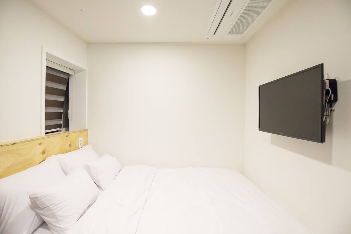 A bedroom with a bed, a window with blinds, a mounted TV, and a ceiling vent.