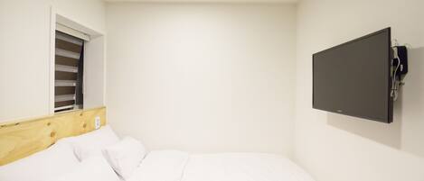 Une chambre à coucher avec un lit, une fenêtre avec des stores, un téléviseur fixé au mur et une bouche d’aération au plafond.