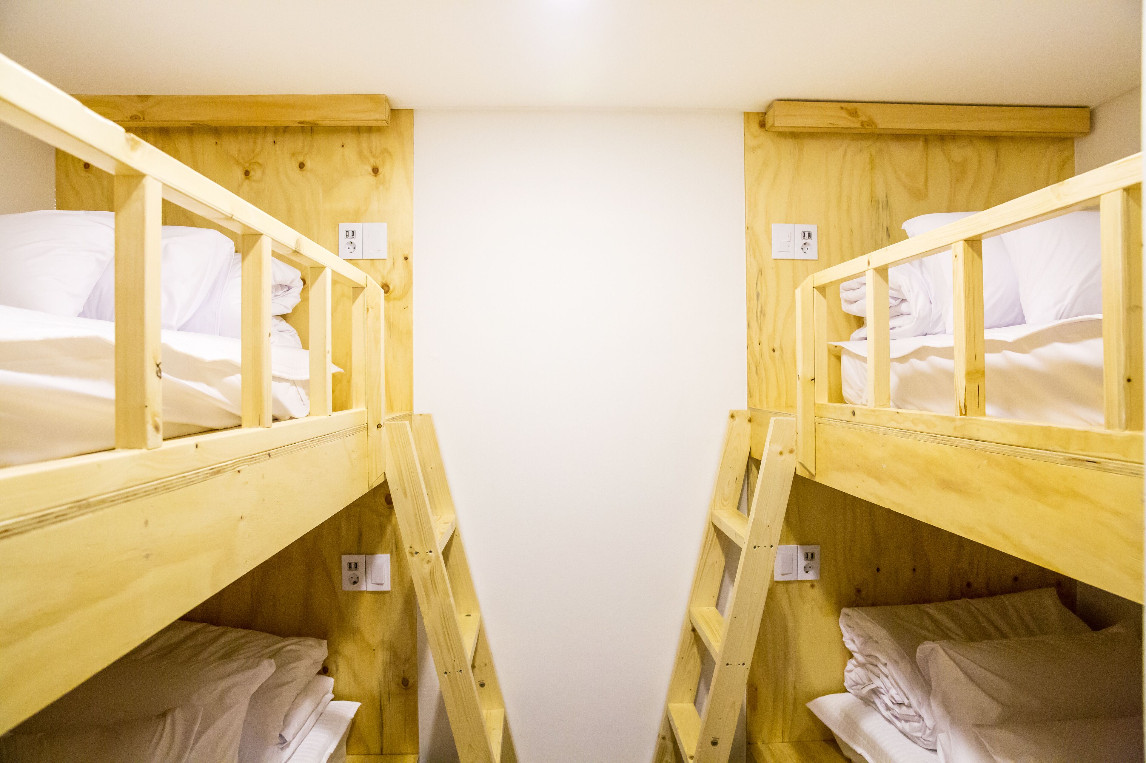 Dos literas con estructura de madera y ropa de cama blanca, dispuestas en una habitación estrecha con una pared blanca lisa.