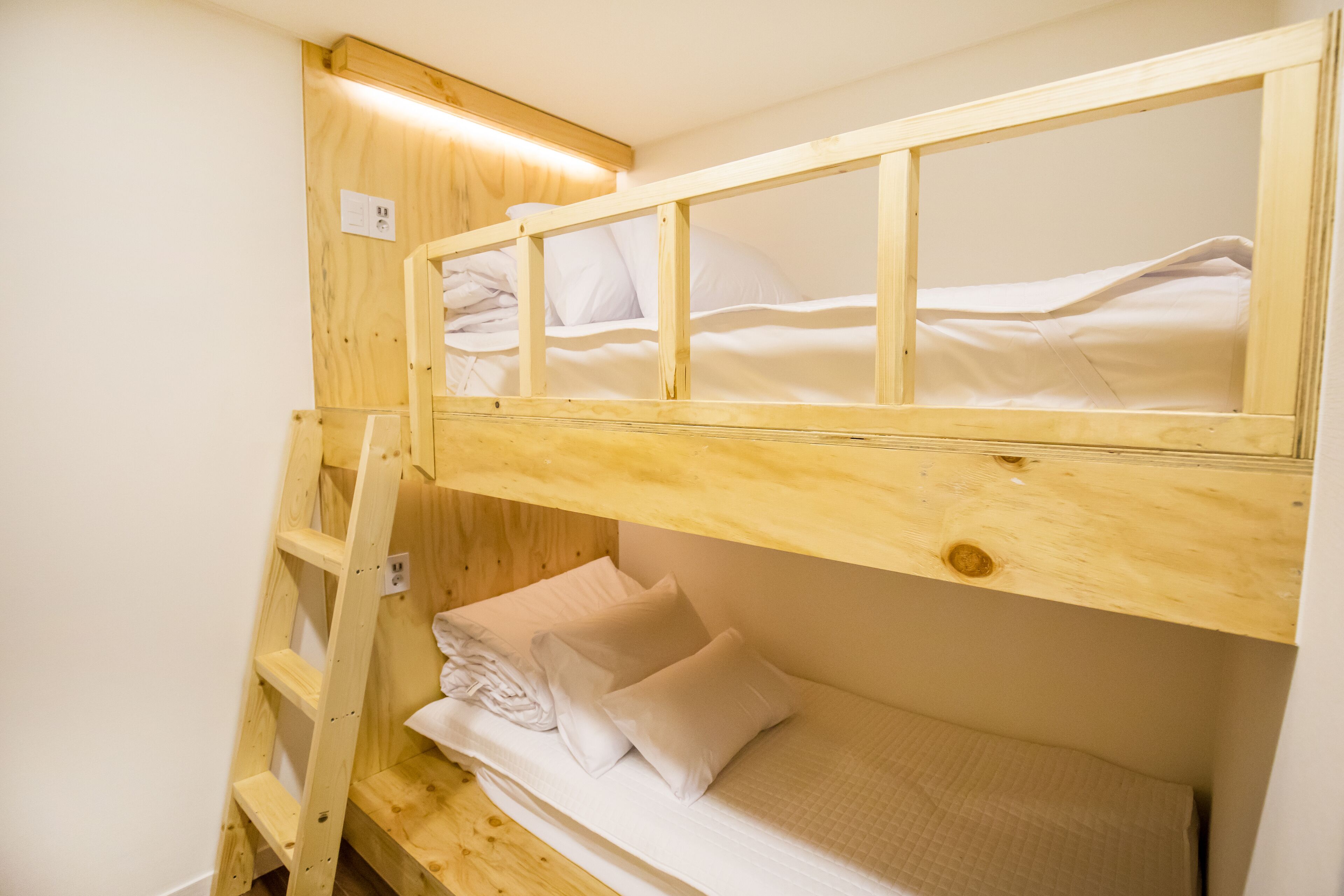 A bunk bed with a wooden ladder, a lower bunk with a mattress and pillows, and a headboard with a light fixture.