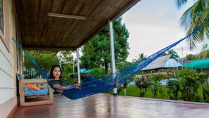 A person relaxing in a hammock on a covered porch with a wooden ceiling, surrounded by greenery and a pool.