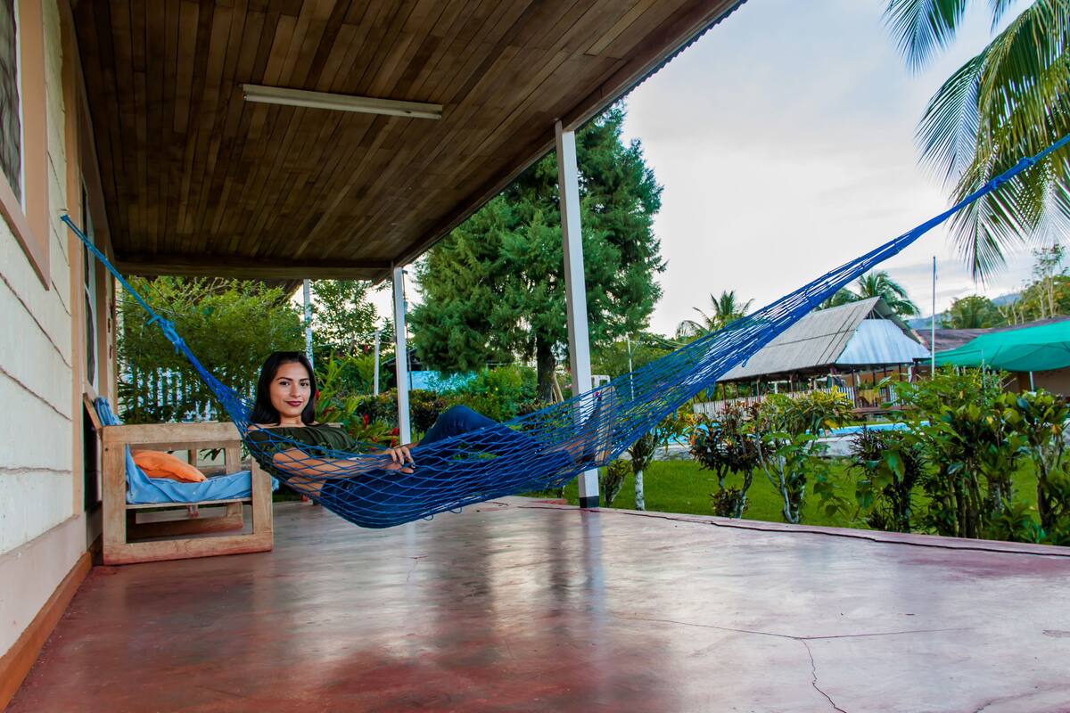 A person relaxing in a hammock on a covered porch with a wooden ceiling, surrounded by greenery and a pool.