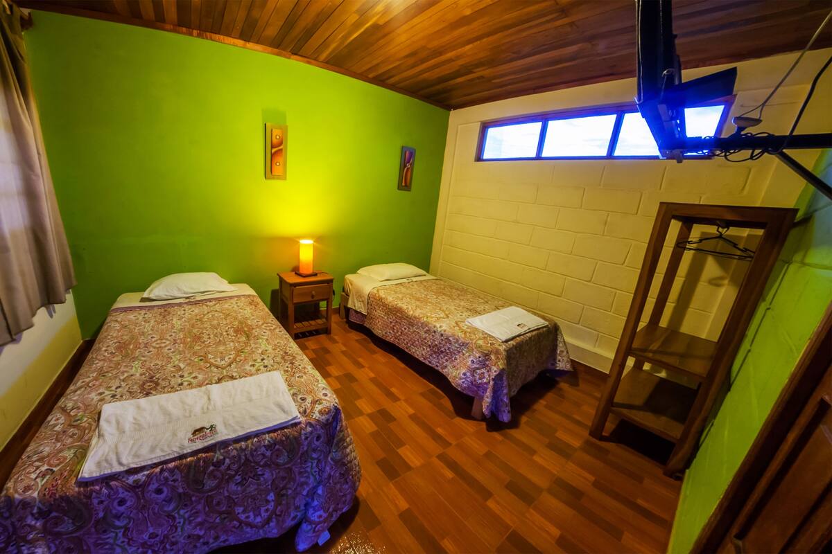 A room with two beds, a wooden ceiling, a green wall, a window, and a hanging light fixture.