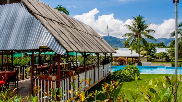 A restaurant with outdoor seating overlooking a pool, surrounded by greenery and palm trees.