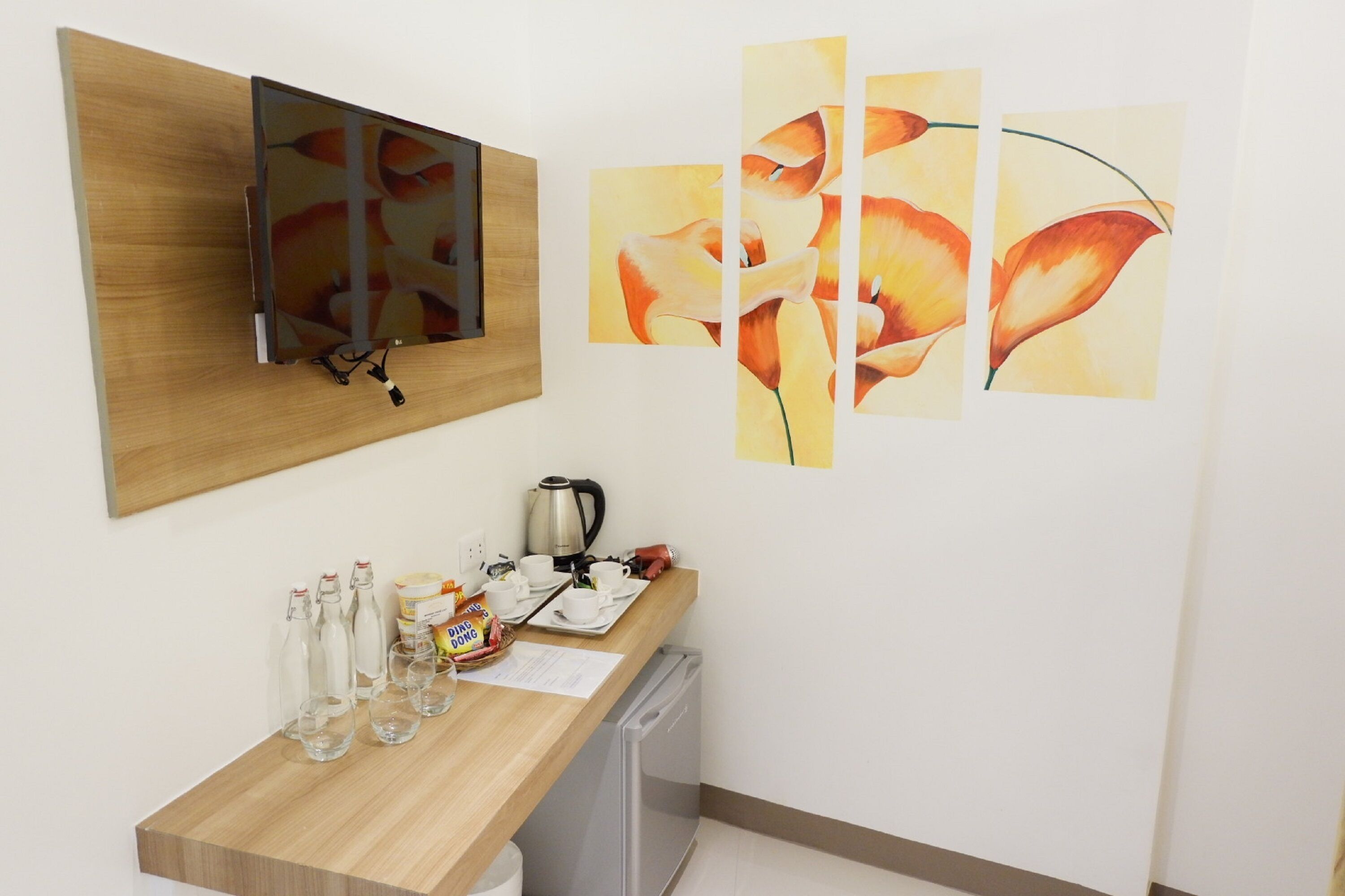 Une chambre d’hôtel avec un bureau en bois, une bouilloire, des tasses et un réfrigérateur. Les murs sont ornés de tableaux et un téléviseur y est fixé.