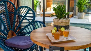 A round wooden table with a tray holding three glasses of amber-colored beverage, garnished with lime slices, and a woven basket with a green plant. Blue wicker chairs with cushions are positioned around the table. - The Akasha Seminyak Bali Boutique Hotel (Keroboken Kelod)