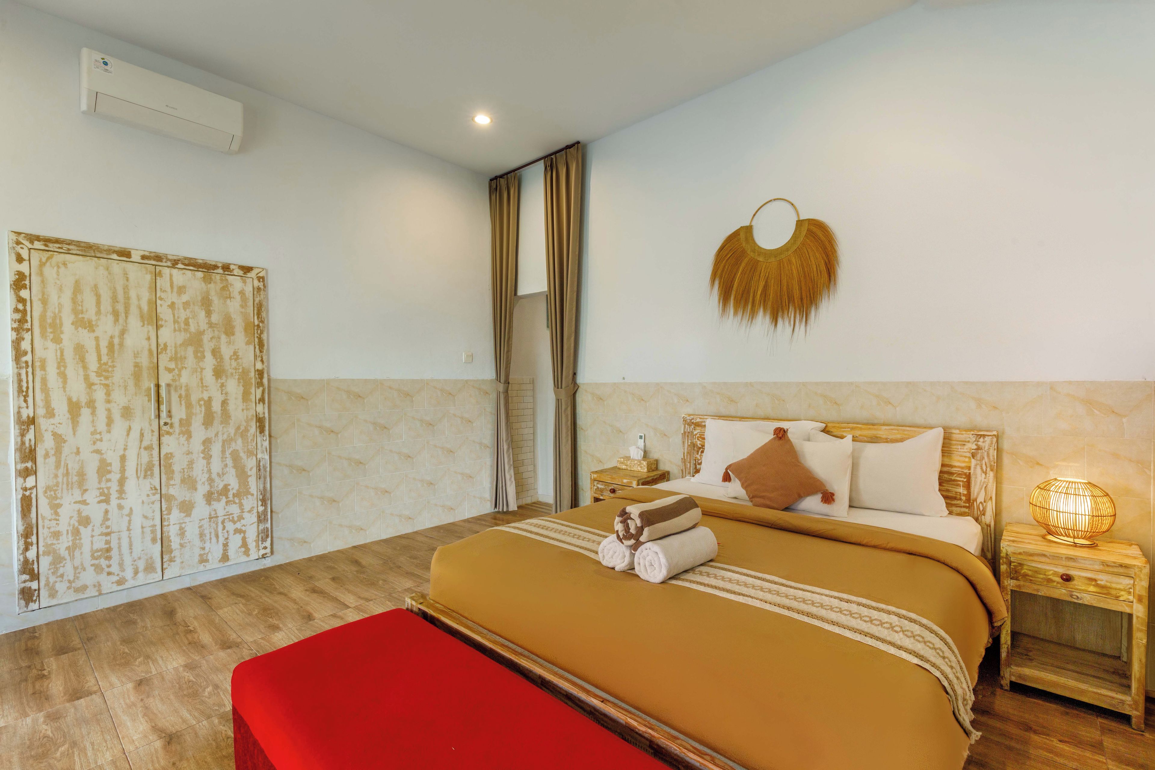A hotel room with a bed, a red bench, a nightstand, a wall-mounted lamp, and a curtain.