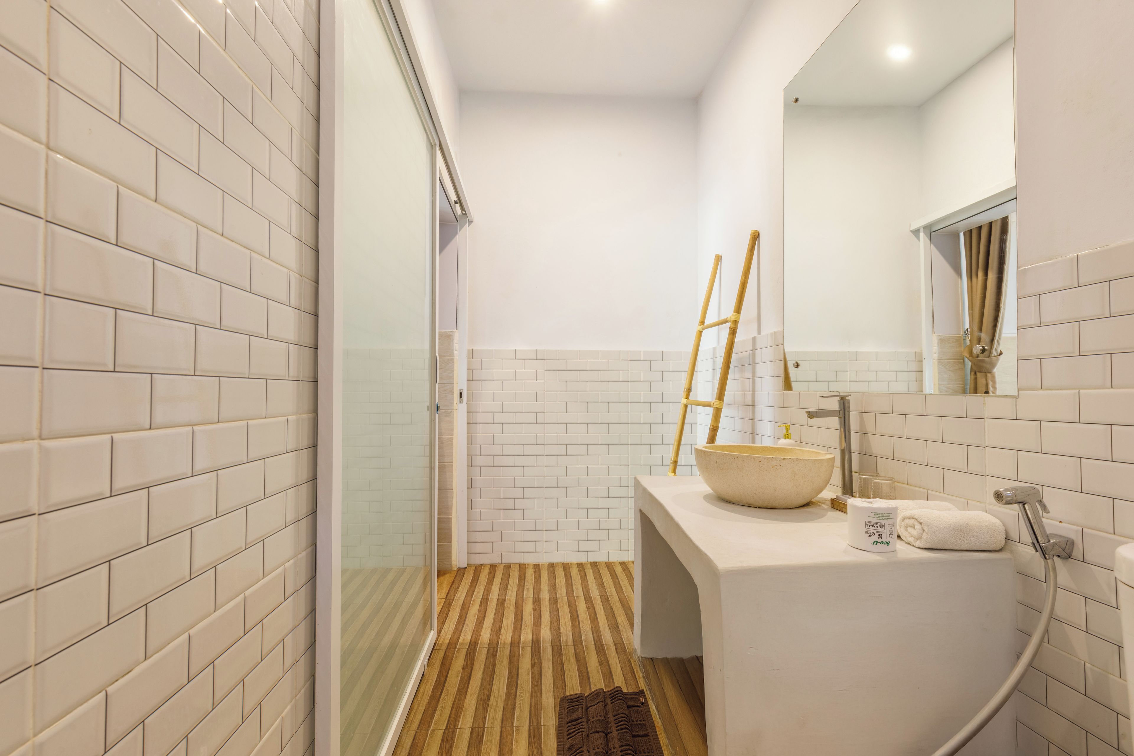 Un baño con paredes de azulejos blancos, suelo de madera, un espejo grande, un lavabo, una toalla y una zona de ducha.