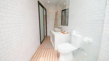Un baño con paredes de ladrillo blanco, suelo de madera, un inodoro, un lavamanos y un espejo.