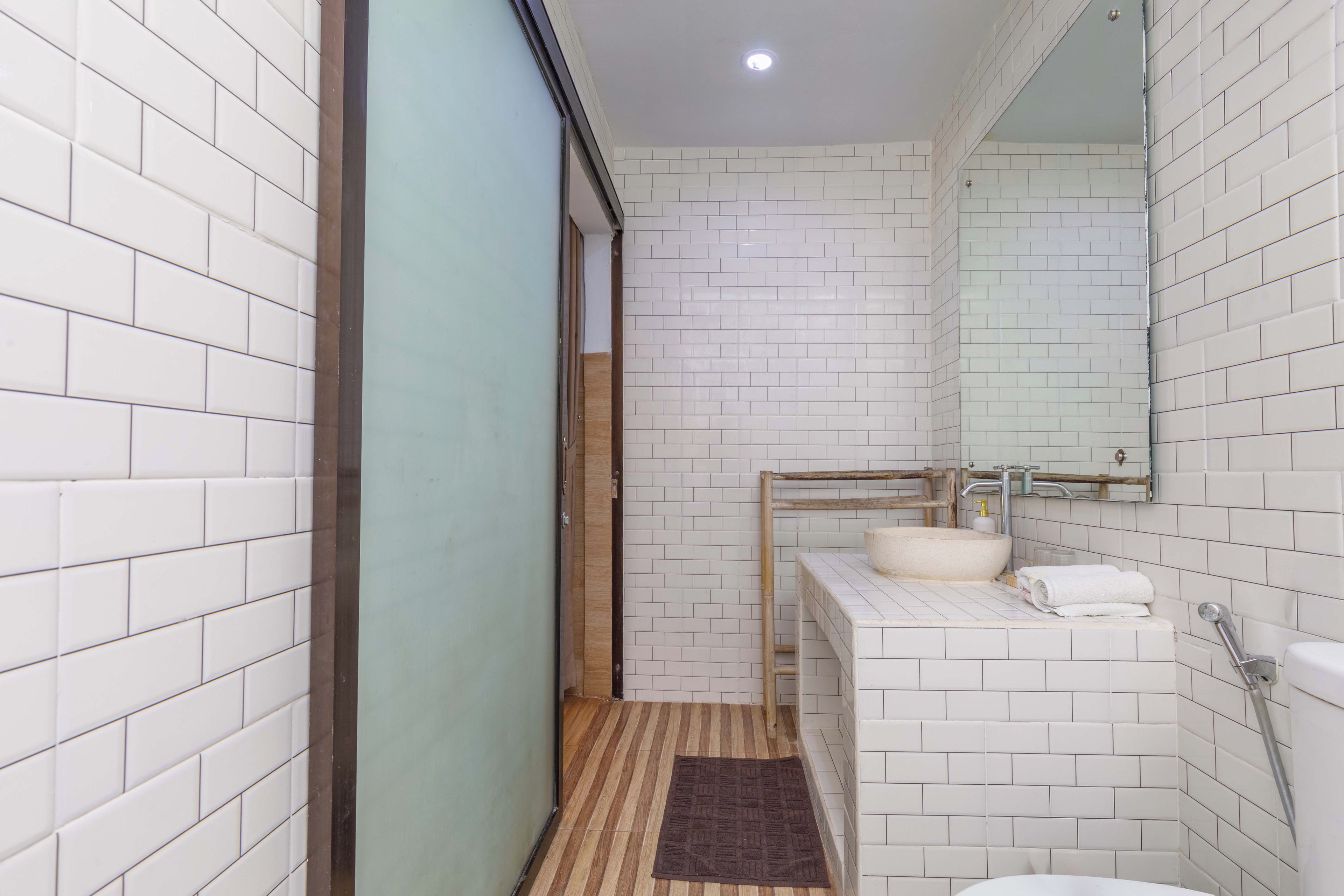 A bathroom with white tiled walls, a wooden door, a sink with a mirror, a shower, and a brown rug.