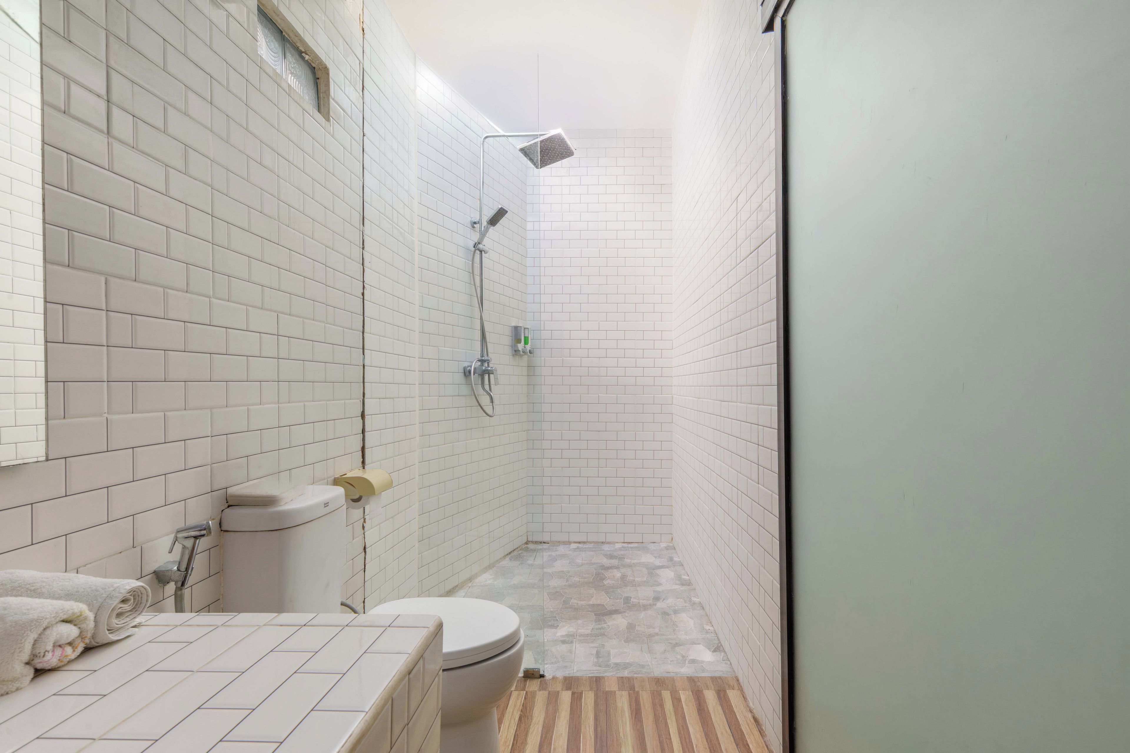 A bathroom with white tiled walls, a toilet, a shower, and a wooden floor.