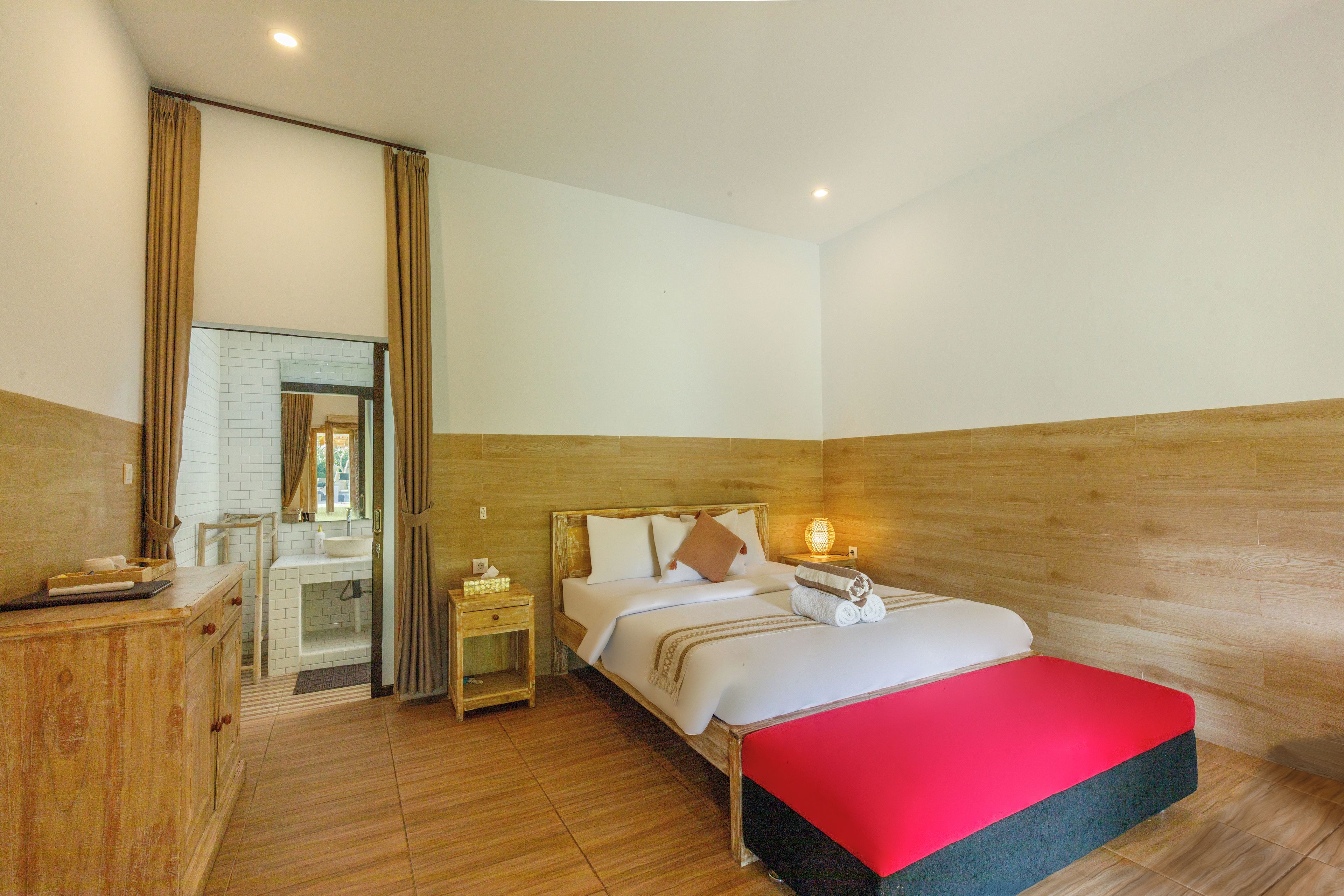A hotel room with a wooden headboard, a bed with white bedding, a red bench, a nightstand with a lamp, and a wooden dresser.
