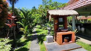 A pathway leading to a thatched-roof structure with a statue, surrounded by lush greenery and a clear blue sky. - Agung Wiwin Homestay (Munggu)