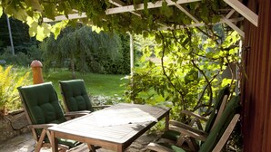 Patio esterno con tavolo in legno e sedie imbottite verdi, circondato da vegetazione rigogliosa e un pergolato.