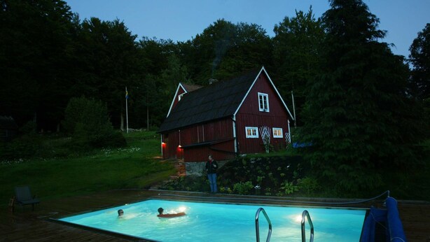 Una piscina con un pontile in legno, una casa rossa sullo sfondo e una persona in piedi vicino alla piscina.