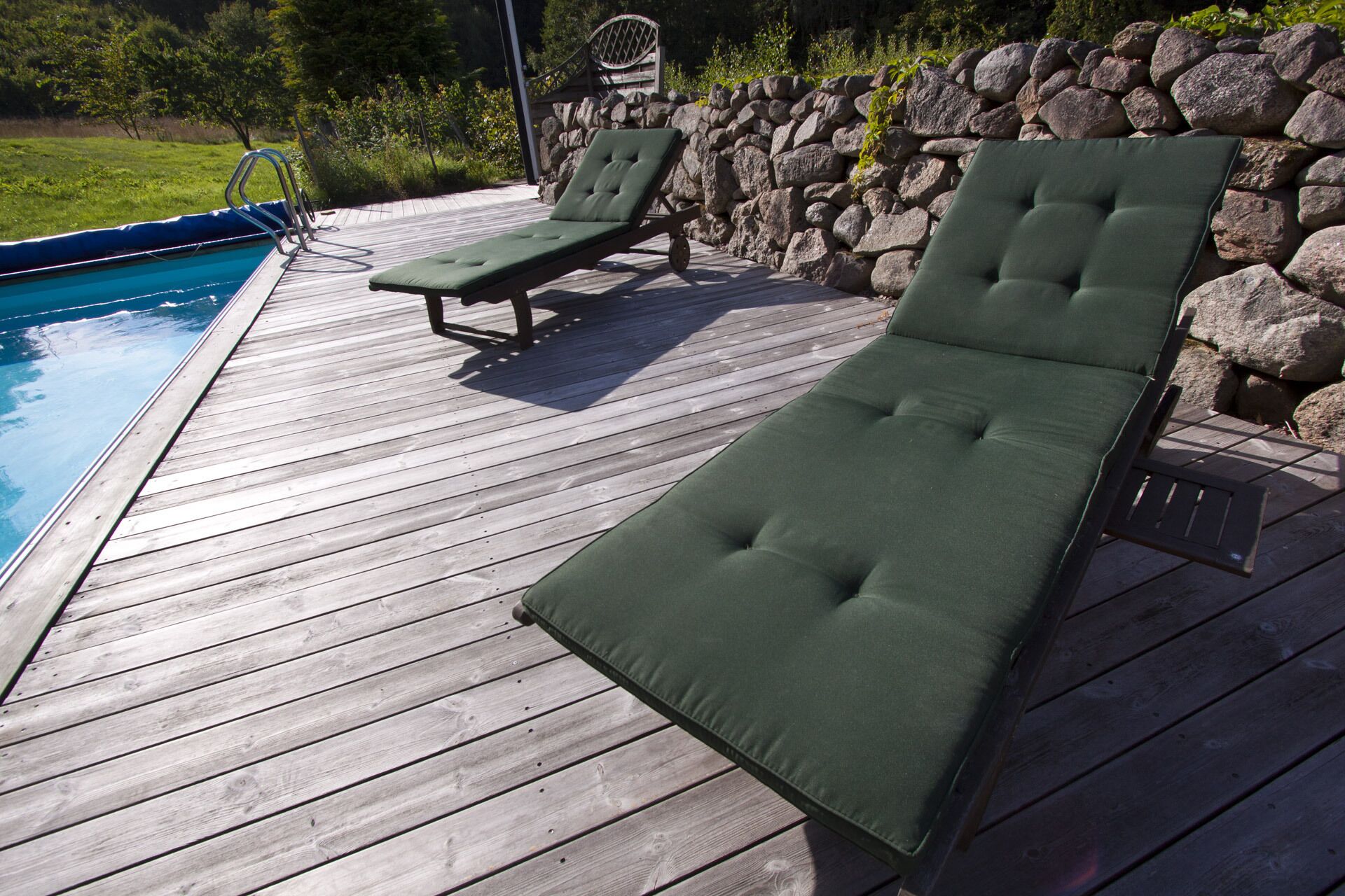 A wooden deck with two green cushioned lounge chairs and a stone wall.