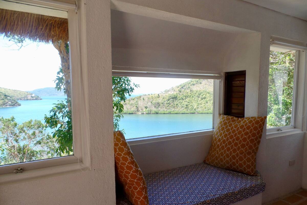 Ein Zimmer mit integriertem Sitzplatz am Fenster mit Seeblick, einem Reetdach und Blick auf Hügel.