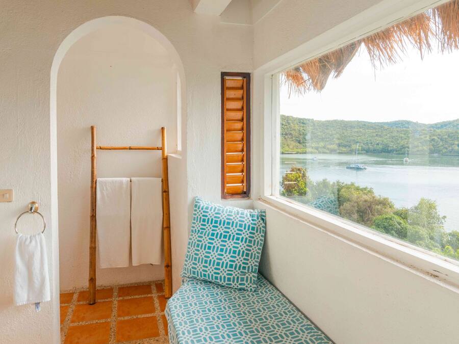 Une salle de bain avec un plafond en bambou, une fenêtre donnant sur un lac, un porte-serviettes et un banc à motifs.