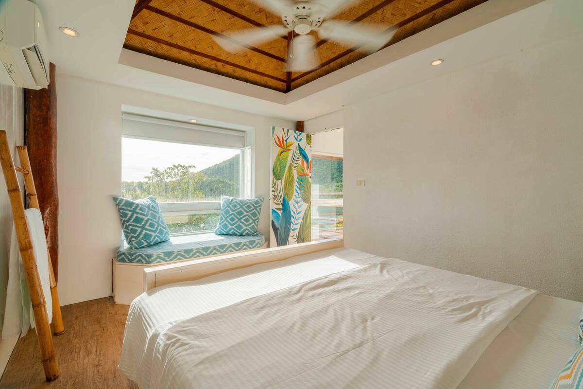 Ein Schlafzimmer mit Bett, Fensterbank mit Kissen, Deckenventilator und Blick ins Grüne.