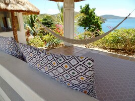 Silla hamaca con cojines estampados, techo de paja y vista a un cuerpo de agua y colinas.