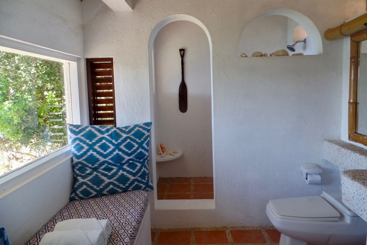 Ein Badezimmer mit Fenster, Dusche, Toilette und einer Sitzbank mit Muster.