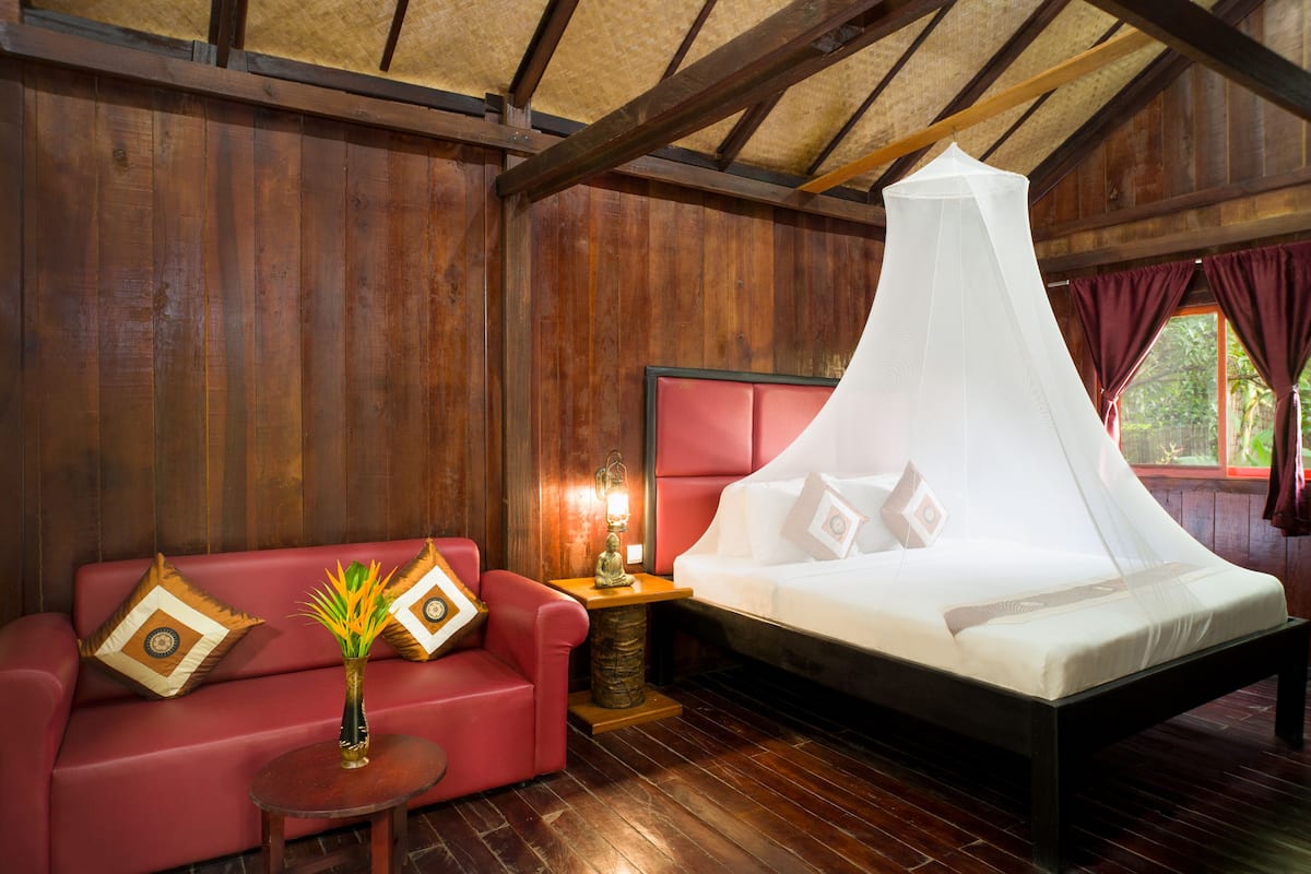 A room with a bed, a red sofa, a small table with a vase, and wooden walls.